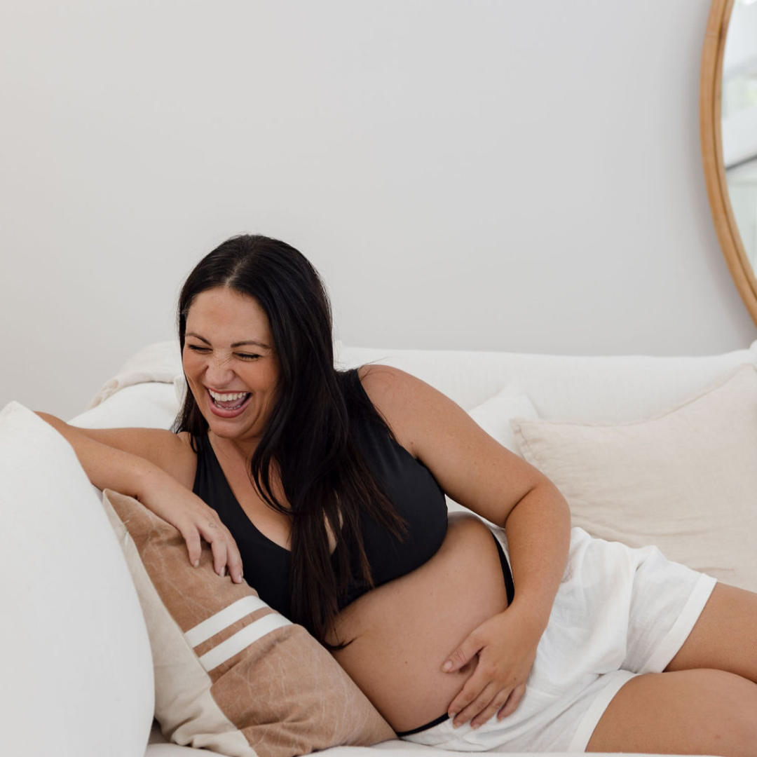 A mum-to-be reclines sideways on a white couch. She is holding her pregnant stomach and laughing, excited about pregnancy.