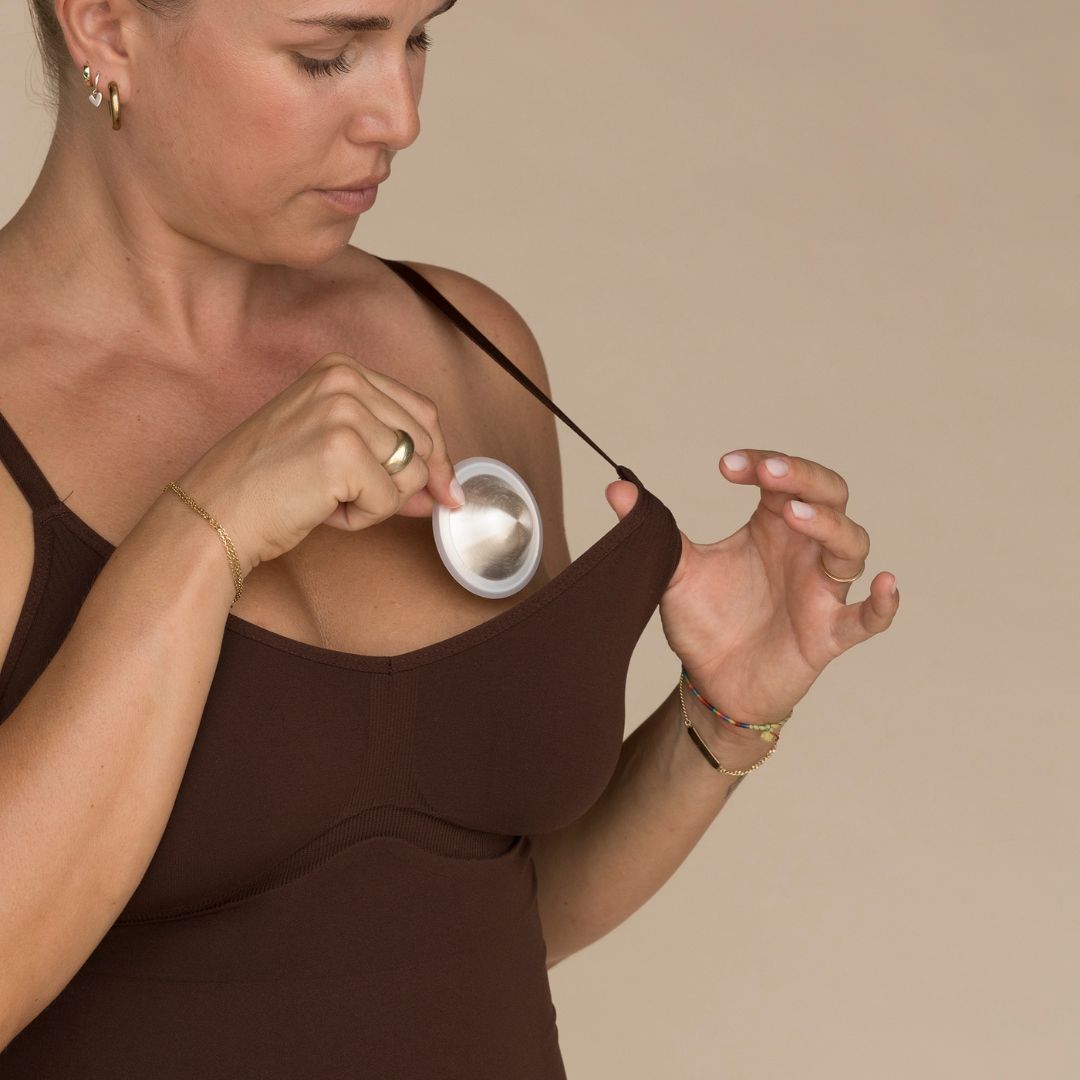 A woman in a brown tank top examines fine Mammae silver nipple cups, pulling them away from the neckline of her top with a focused expression.