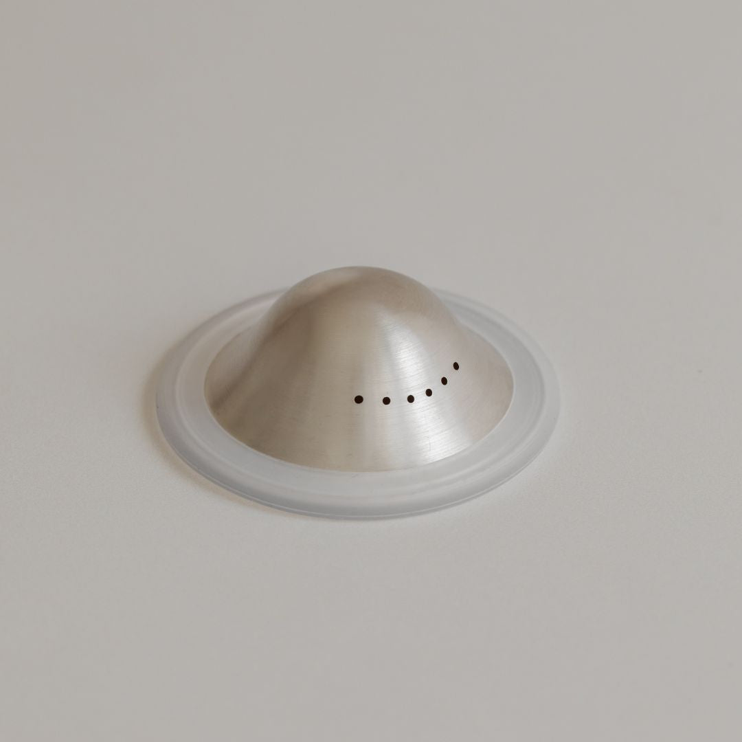 A Mammae silver nipple cups with vent holes, set on a circular transparent base, against a light grey background.