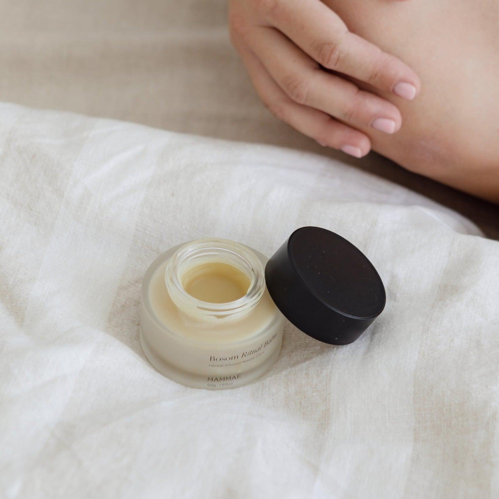 A breastfeeding mother using Mammae bosom ritual balm for postpartum breast care while sitting on a bed.