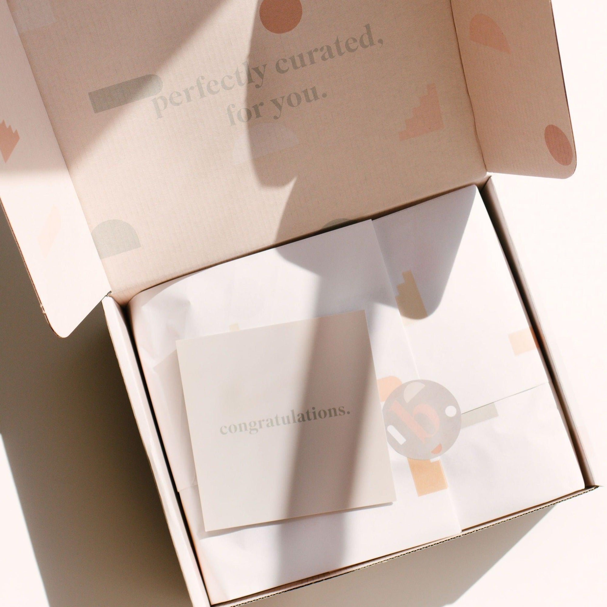 An open cardboard box reveals a neatly wrapped item with a card on top from biglittlethings that reads "congratulations." The inside lid of the box says, "perfectly curated, for you.