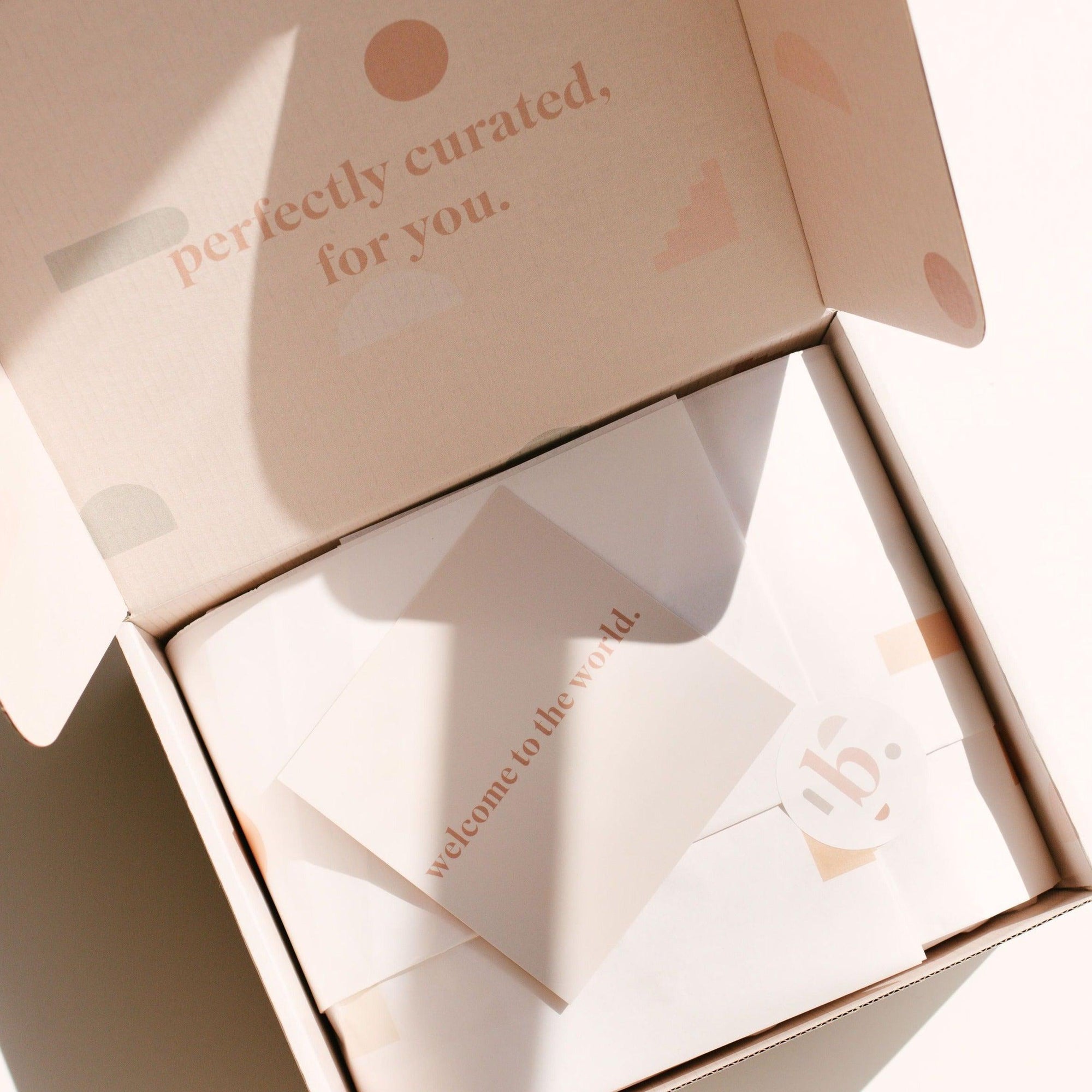 A slightly open cardboard box with text reading "perfectly curated, for you" reveals a gift card titled "welcome to the world" from biglittlethings., accompanied by some folded papers.