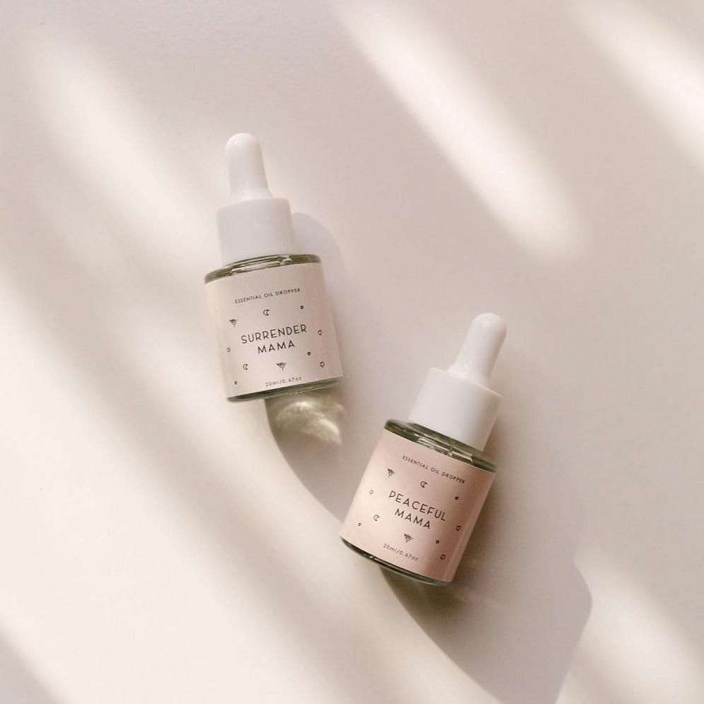 Two small bottles of Surrender Mama Essential Oil Dropper and Peaceful Mama Essential Oil Dropper by Seasons of Mama on a white surface.