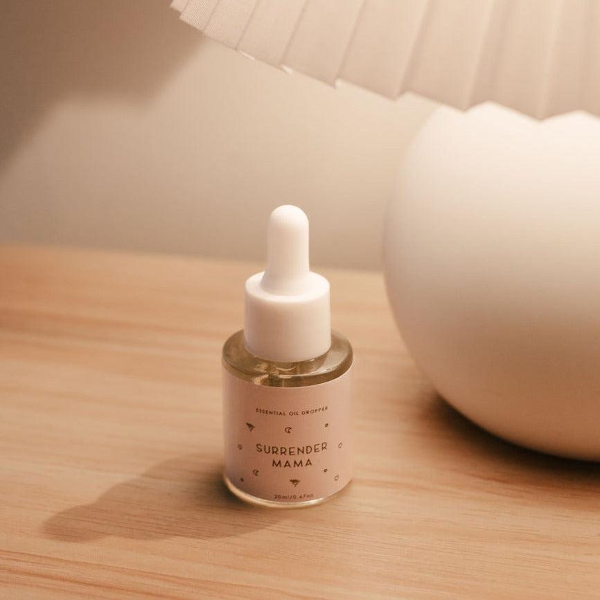A bottle of Seasons of Mama's Surrender Mama Essential Oil Dropper sitting on a table next to a lamp.