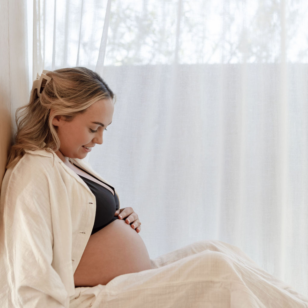 A side profile of a pregnant woman sitting in front of a window with a white linen curtain. She is wearing linen maternity wear and a black maternity bra, and holding her pregnant bump lovingly. She looks relaxed and ready for a calm birth process.