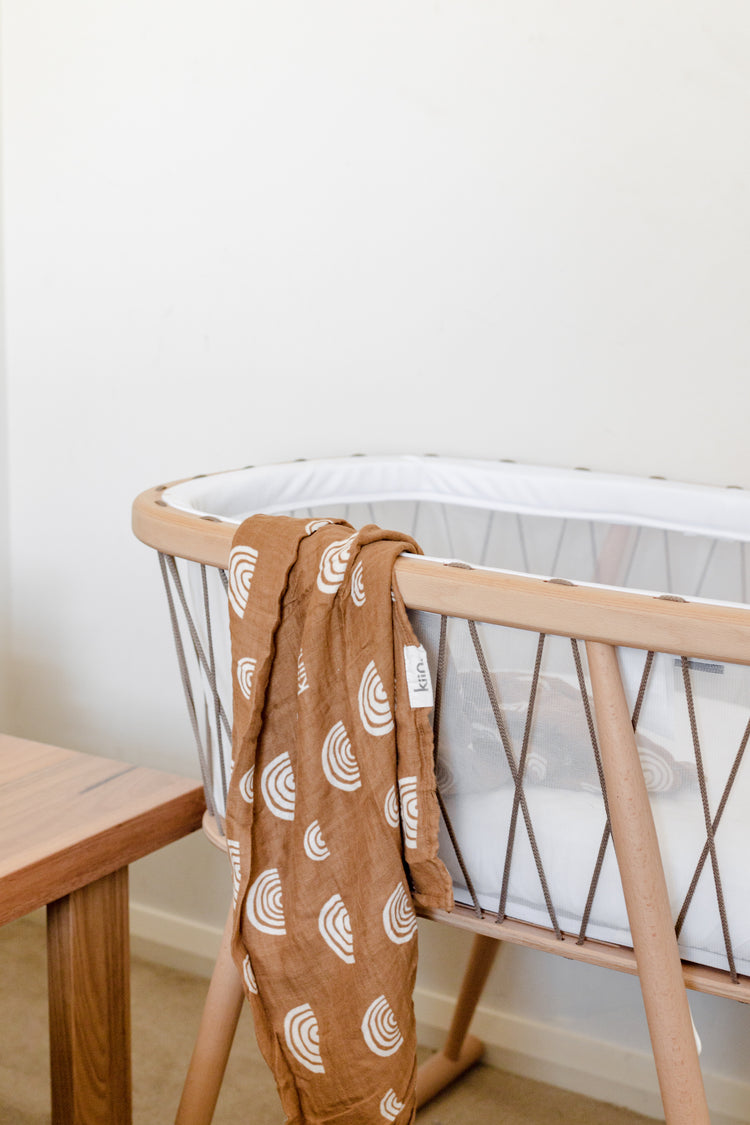 Baby bassinet with a brown patterned blanket draped over the edge, next to a wooden table.