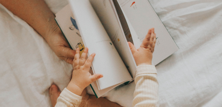 A baby wears a striped onesie and reaches for a book being held by an adult, while sitting on a white bed.