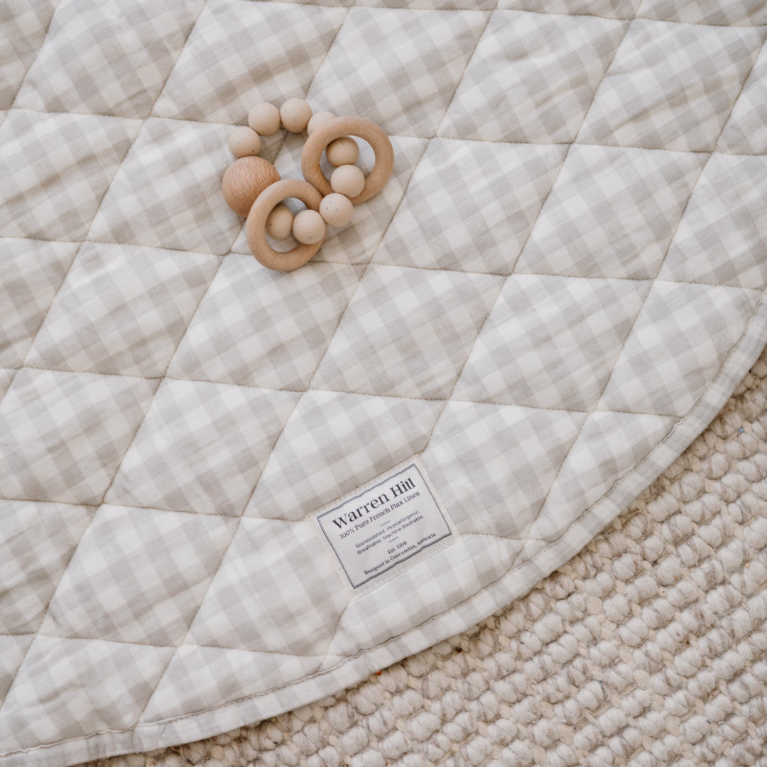 A wooden baby teether rests on a Warren Hill play mat with a gingham pattern, placed over a textured rug.