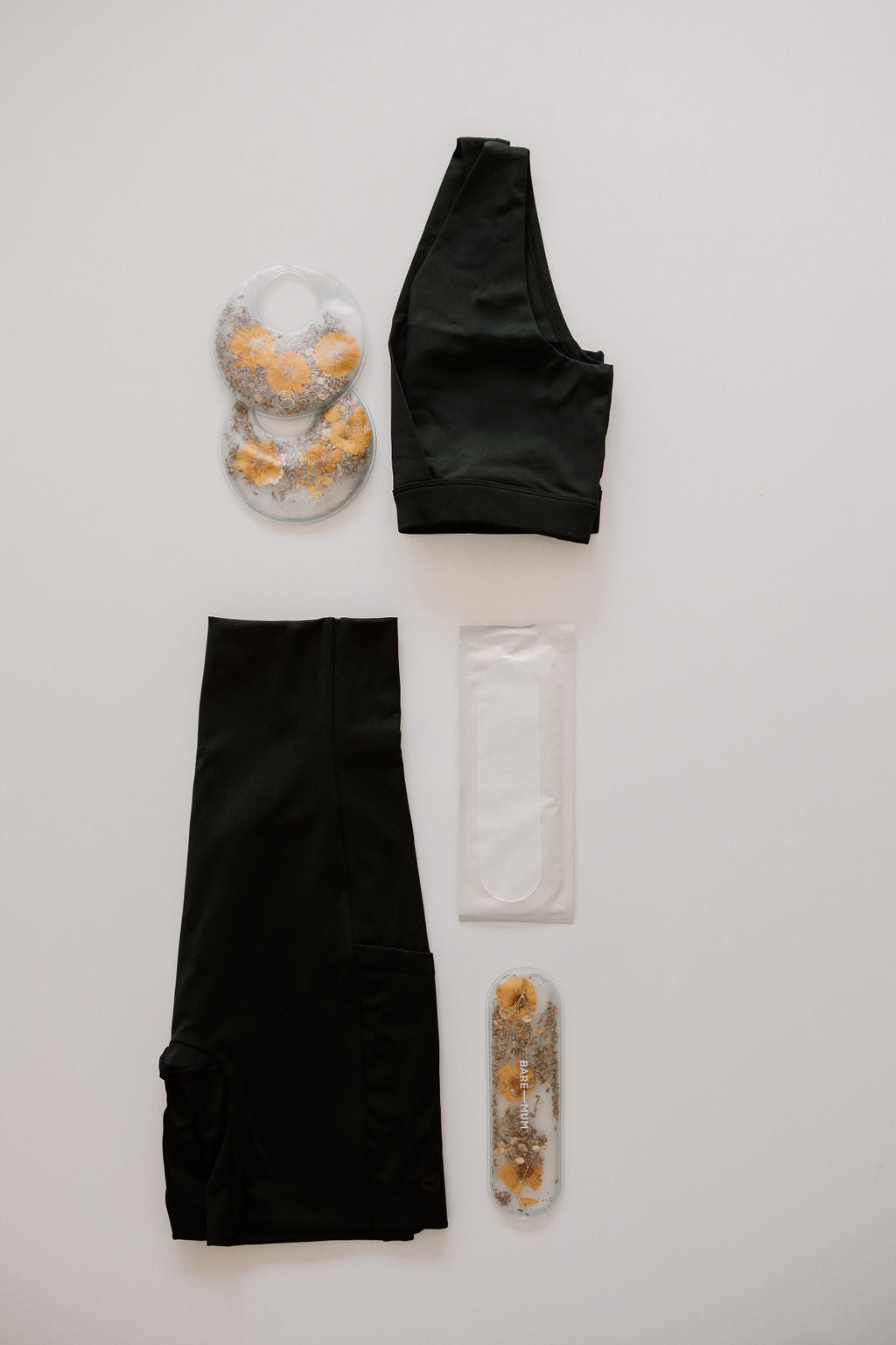 A **Bare Mum bare mum recovery kit for c-section** featuring a black sports bra, black leggings, two gel ice packs with orange spheres, and two absorbent pads is laid out on a white surface.