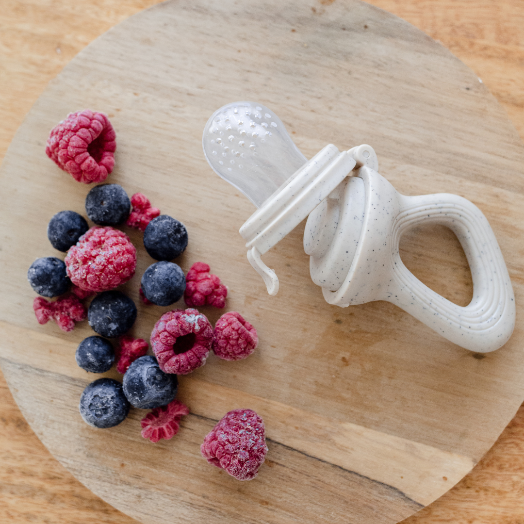A Dove & Dovelet silicone self feeder next to raspberries and blueberries on a wooden board.