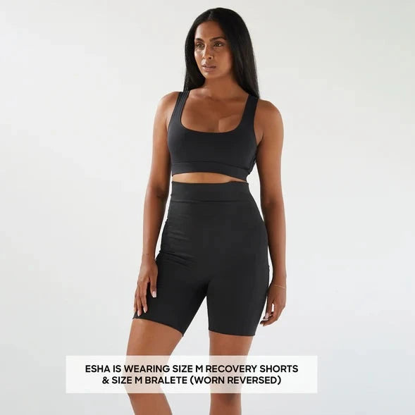 A woman in a black sports bra and Bare Mum Postpartum Recovery Shorts standing against a light background, labeled with her outfit details.