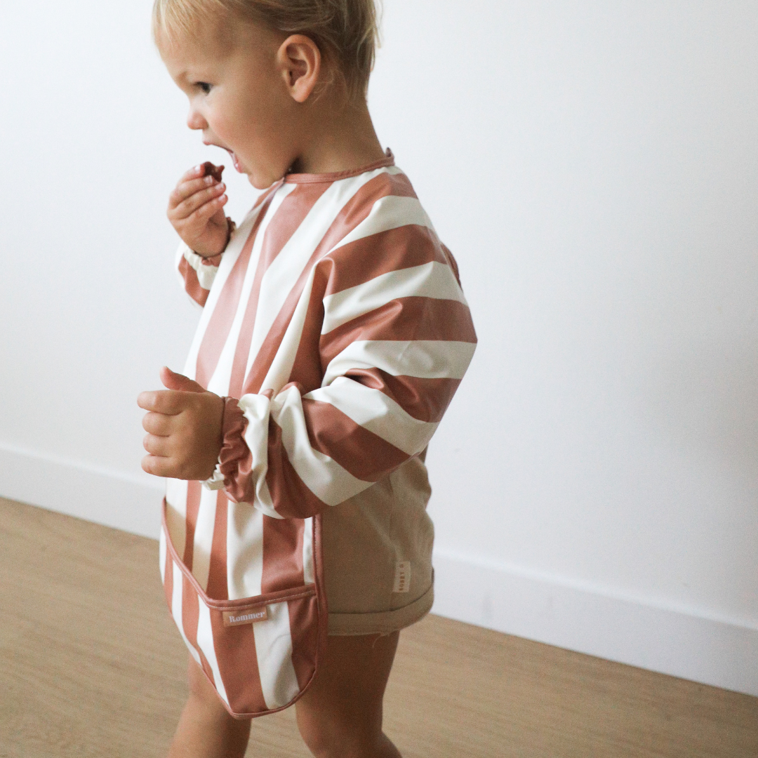A child wearing a Rommer Smock Bib in the cinnamon color variation in a super soft fabric.