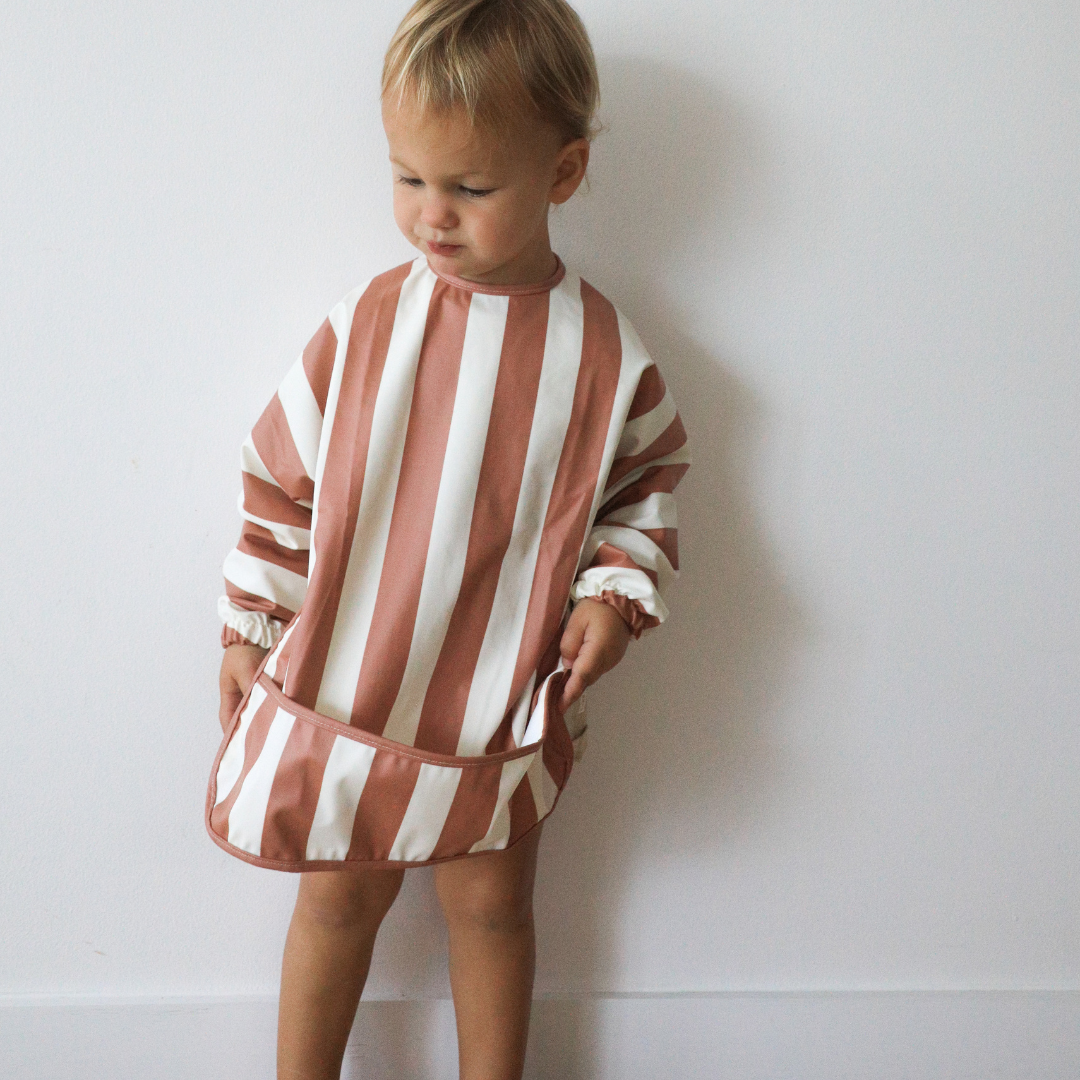 A little boy in a Rommer Smock Bib | cinnamon and white striped dress.