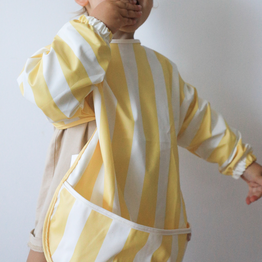 A child wearing a Super Soft fabric, Rommer Smock Bib by Rommer that features striped color variations in yellow and white.