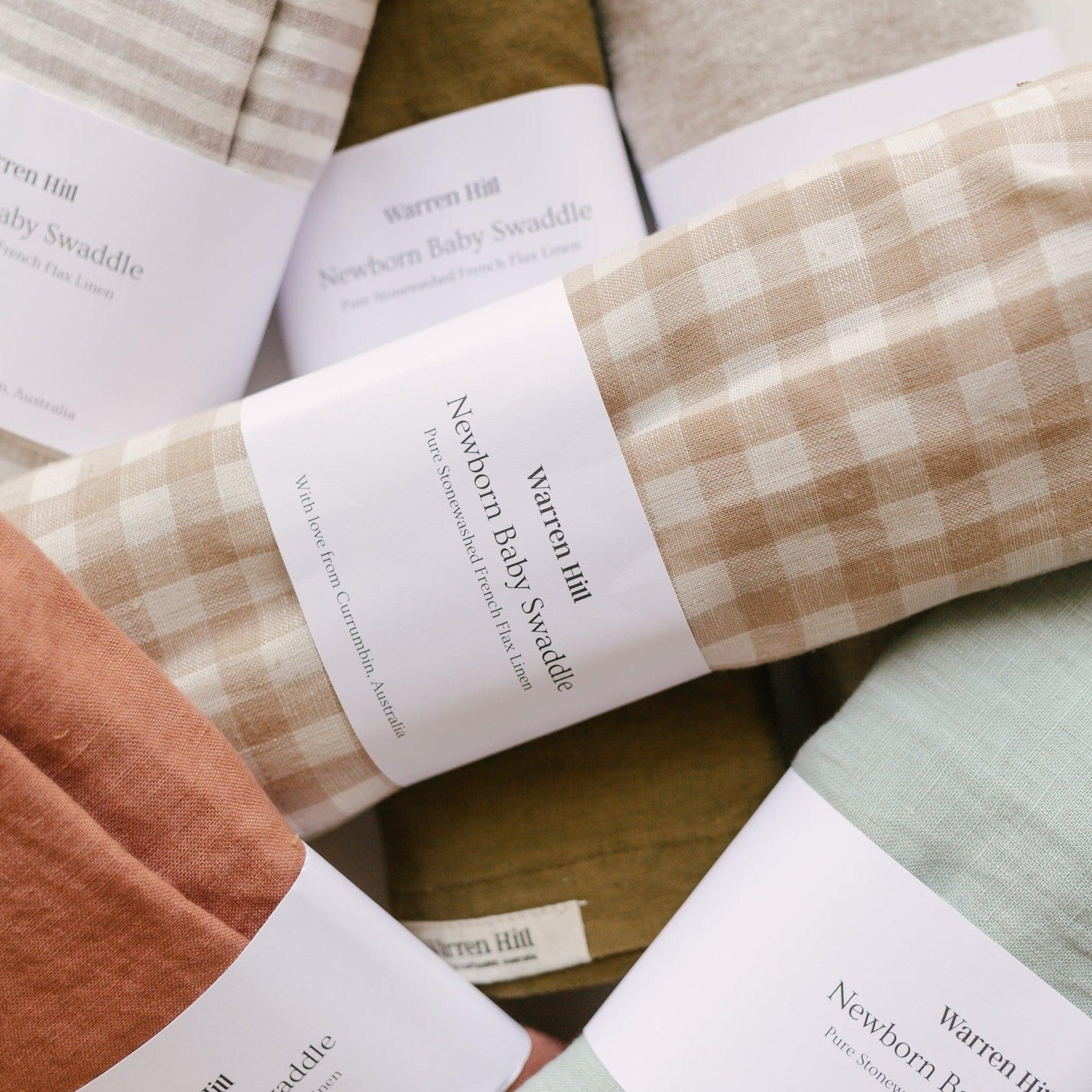 A group of Warren Hill towels with different colors and patterns, including French linen baby swaddle | beige gingham styles.