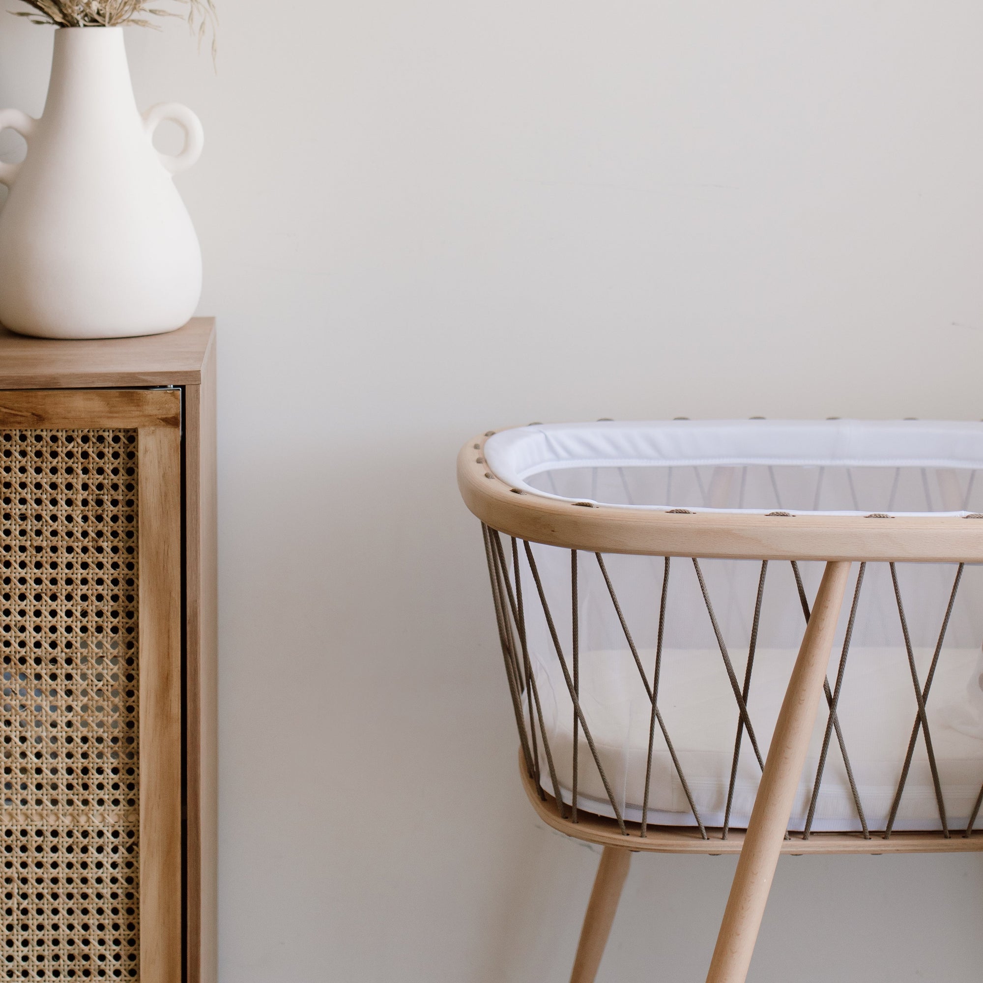 A KUMI bassinet in hazelnut colour by Charlie Crane adjacent to a wooden chest of drawers.