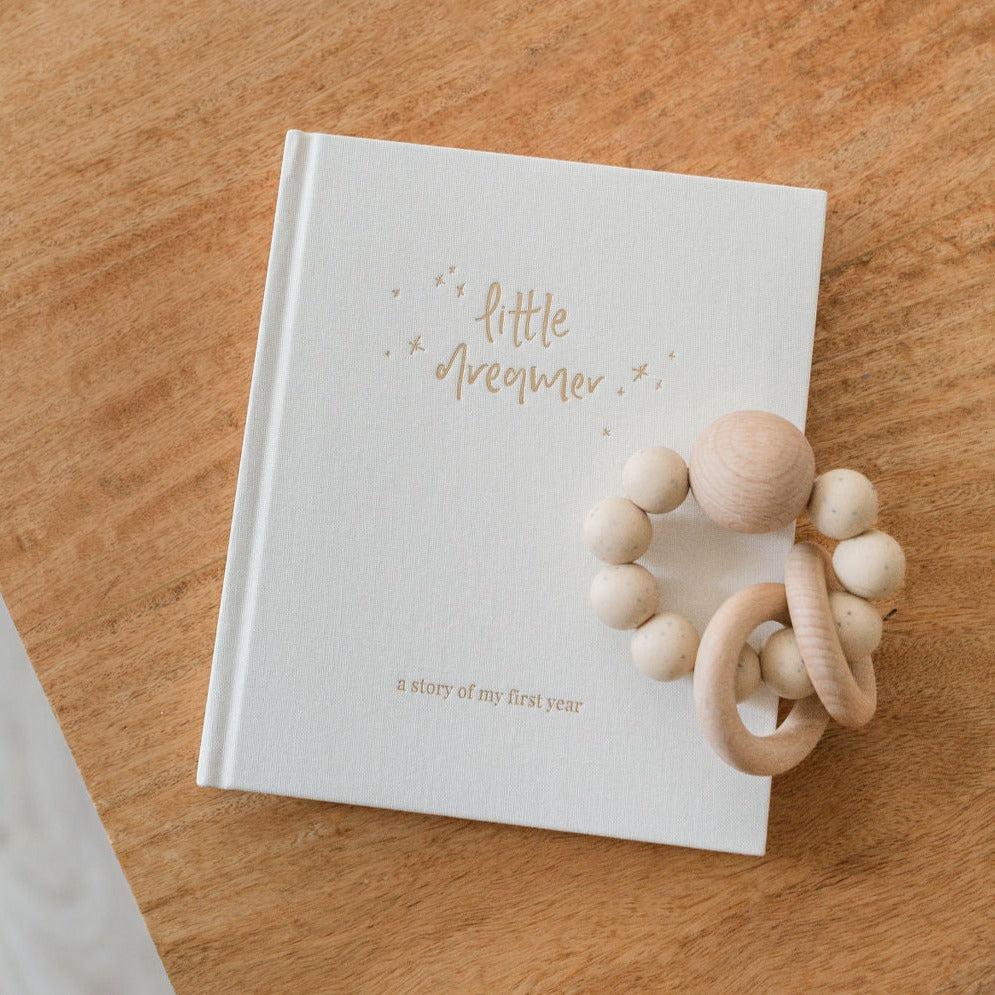 An Emma Kate Co. little dreamer baby journal in cloud on a timber surface with a wooden baby rattle on top of it.