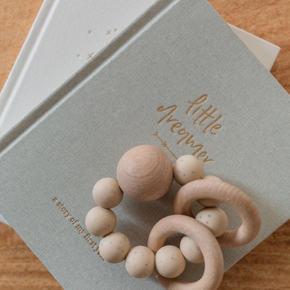 An Emma Kate Co. little dreamer baby journal in sage on a timber surface with a wooden baby rattle on top of it.