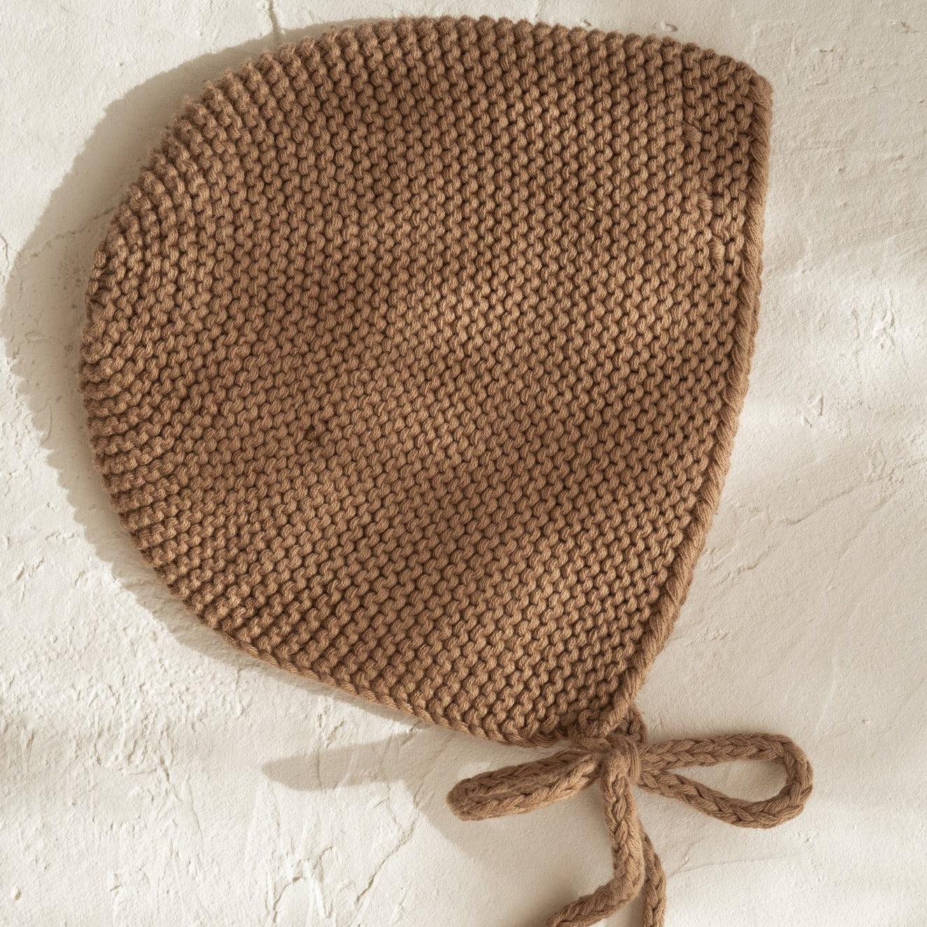 An illoura baby bonnet in chocolate on a white wall.