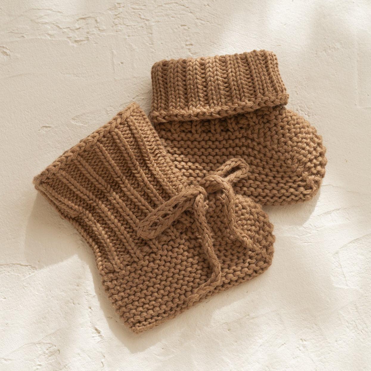 A pair of Illoura the Label organic cotton knitted baby booties in chocolate on a white surface.